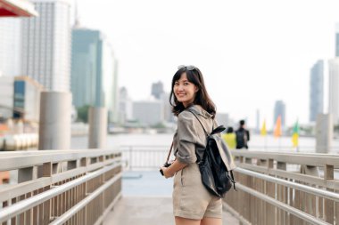 Young Asian woman backpack traveler waiting an express boat at pier on Chao Phraya River in Bangkok. Traveler checking out side streets.