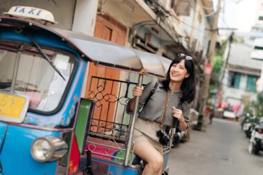 Young Asian woman backpack traveler standing a side of Tuk Tuk taxi on summer vacations at Bangkok, Thailand. Journey trip lifestyle, Asia summer tourism concept.