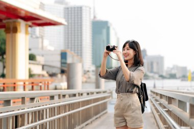 Young Asian woman backpack traveler using a camera in express boat pier on Chao Phraya River in Bangkok. Traveler checking out side streets. clipart