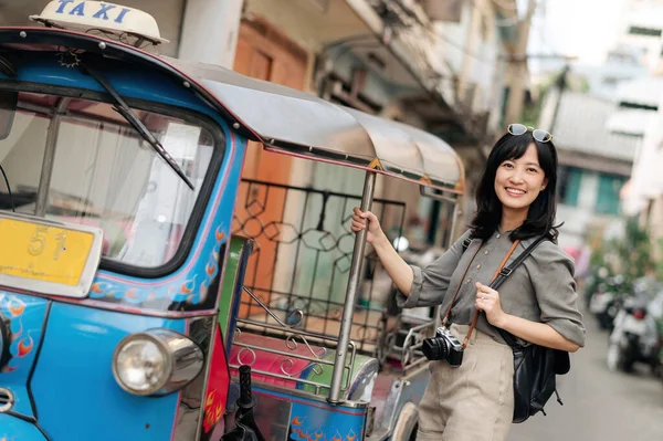 Young Asian woman backpack traveler standing a side of Tuk Tuk taxi on summer vacations at Bangkok, Thailand. Asia summer tourism concept or world travel explorer.