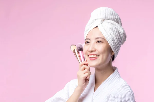 Portrait young happy asian woman with natural make up face holding cosmetic skin powder blusher isolated on pink background. Female apply skincare brush treatment. beauty product, cosmetology concept.