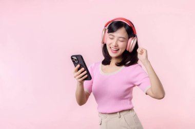 Portre genç, neşeli Asyalı kadın pembeye karşı akıllı telefon müzik uygulaması ile ses dinlemeyi seviyor. Kulaklıklı, mutlu, gülümseyen bir kadın. Ses, eğlence, yaşam tarzı, teknoloji konsepti