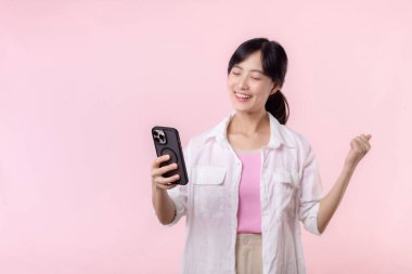 Güzel Asyalı bir kadın başarı gösterirken zafer el hareketi yaparken aynı zamanda akıllı telefondan pembe arka planda harika haberler alıyor. Mutlu teknoloji, çevrimiçi uygulama, cep telefonu reklam kavramı