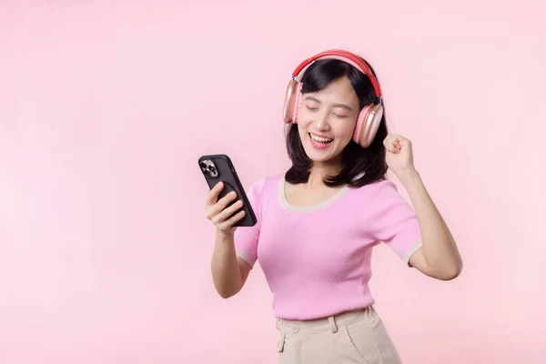 若い陽気なアジアの女性の肖像画は ピンクに対するスマートフォンの音楽アプリケーションによってオーディオを聞いてお楽しみください ヘッドフォンで幸せな笑顔の女性の人 レジャー ライフスタイル テクノロジーの概念 — ストック写真