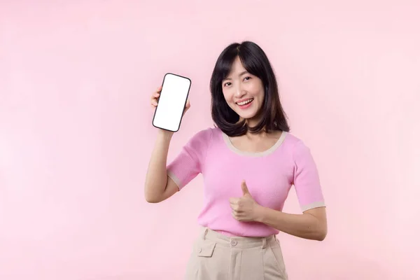 在粉红的画室背景下 年轻美丽的亚洲女人带着手指手势 对着手机 开心地微笑着 展示着智能手机屏幕 — 图库照片
