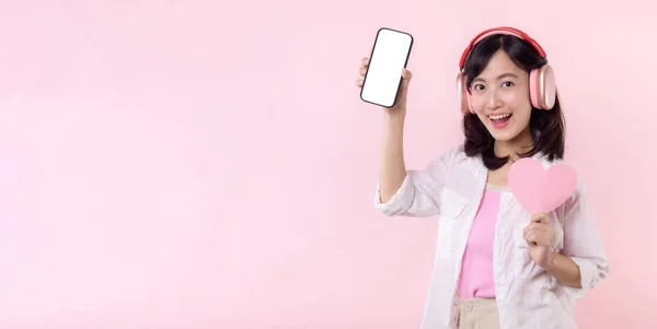 快乐的笑着的亚洲女人 手牵着心和耳机 展示着空白屏幕手机或在粉色工作室背景下孤立的新智能手机音乐应用程序广告模型 — 图库照片