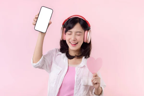 携帯電話や新しいスマートフォンの音楽アプリケーション広告モックアップピンクのスタジオの背景に隔離された白い画面の携帯電話を示す手の心とイヤフォンを持つ幸せな笑顔陽気なアジアの女性 — ストック写真