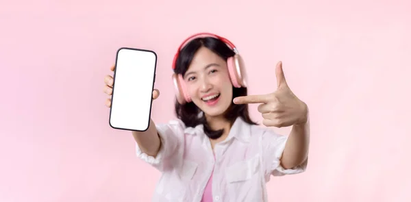 快乐的笑着的亚洲女人 带着无线耳机 展示空白屏幕手机或新的智能手机音乐应用程序广告模型 在粉红的工作室背景下被隔离 — 图库照片