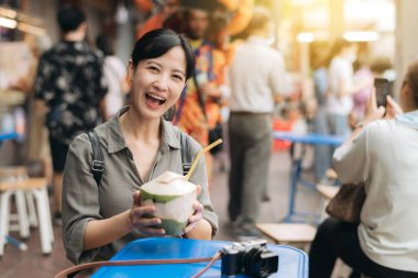 Happy young Asian woman backpack traveler drinking a coconut juice at China town street food market in Bangkok, Thailand.