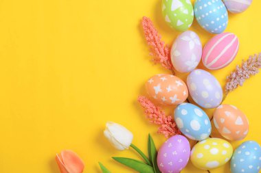 Mutlu Paskalya bayramı tebrik kartı konsepti. Sarı arka planda renkli Paskalya yumurtaları ve bahar çiçekleri. Üst görünüm, düz görünüm, kopyalama alanı.