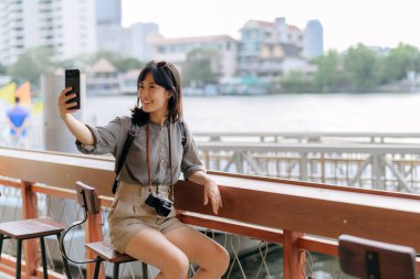 Young Asian woman backpack traveler using mobile phone in express boat pier on Chao Phraya River in Bangkok. Traveler checking out side streets.