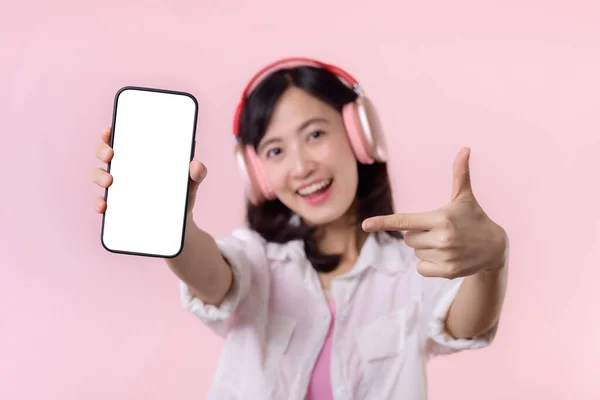ピンクのスタジオの背景に隔離された白い画面の携帯電話や新しいスマートフォンの音楽アプリケーション広告モックアップを示す無線イヤフォンを持つ幸せな笑顔陽気なアジアの女性 — ストック写真