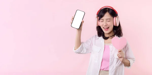 快乐的笑着的亚洲女人 手牵着心和耳机 展示着空白屏幕手机或在粉色工作室背景下孤立的新智能手机音乐应用程序广告模型 — 图库照片