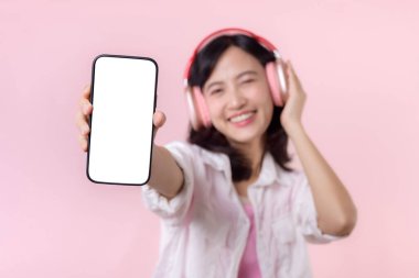 Kablosuz kulaklıklı, mutlu gülümseyen Asyalı kadın boş ekran cep telefonu ya da yeni akıllı telefon müzik uygulaması reklamı gösteriyordu. Pembe stüdyo arka planında izole edilmiş..
