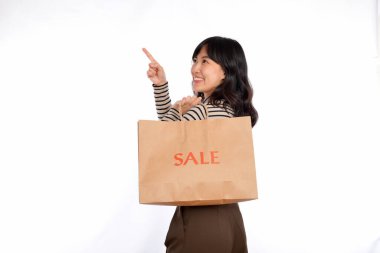 Young Asian woman on sweater cloth pointing up to copy space and looking at camera with smile face and sale shopping paper bag isolated on white background