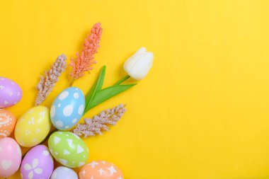 Mutlu Paskalya bayramı tebrik kartı konsepti. Sarı arka planda renkli Paskalya yumurtaları ve bahar çiçekleri. Üst görünüm, düz görünüm, kopyalama alanı.