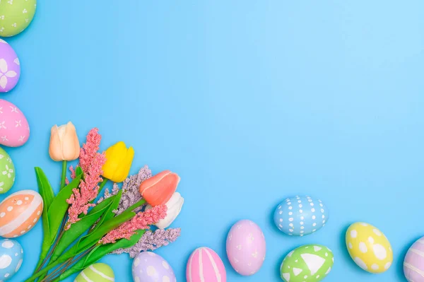 stock image Happy Easter holiday greeting card concept. Colorful Easter Eggs and spring flowers on blue background. Top view, flat lay, copy space.