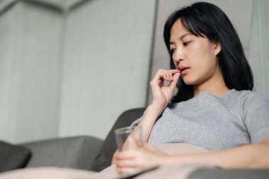 Asyalı genç bir kadın kanepede yatarken ilaç alıyor.