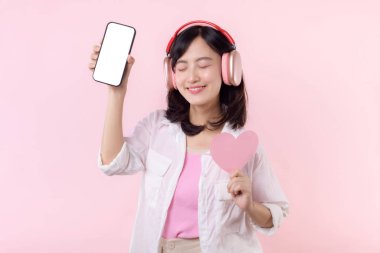 Elinde kalp ve kulaklıkla gülümseyen mutlu Asyalı kadın boş ekran cep telefonu ya da yeni akıllı telefon müzik uygulaması reklamı gösteren pembe stüdyo arka planında izole edilmiş..