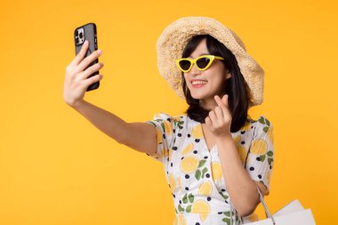 Çekici, mutlu Asyalı bir kadın şık bir bahar elbisesi, şapkası, güneş gözlüğü modası ve sarı arka planda izole edilmiş kese kağıdıyla akıllı telefon kullanıyor. Yaz mevsiminde alışveriş konsepti.