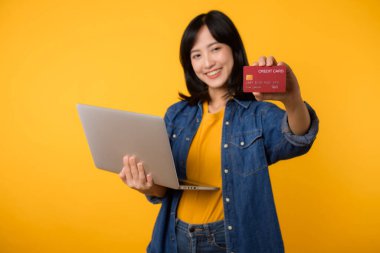 Asya kökenli mutlu genç bir kadın dizüstü bilgisayar kullanarak sarı tişört giyer kredi kartı alışverişi yapar sarı arka planda izole edilmiş banka kartı siparişi verir..