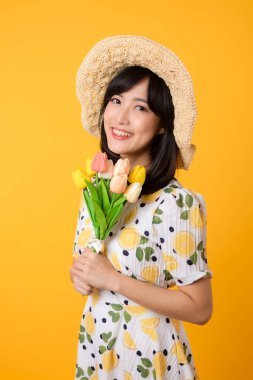 Stüdyo portresi güzel Asyalı genç kadın mutlu bir gülümseme takınıyor ve sarı stüdyo arka planında lale çiçeği tutan bir şapkası var. Kişisel tatil konsepti.