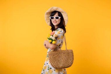 Stüdyo portresi oldukça genç Asyalı mutlu bir gülümseme. İlkbahar modası, güneş gözlüğü, dokuma çanta ve sarı stüdyo arka planında lale çiçeği tutuyor. Kişisel tatil konsepti.