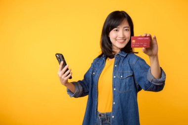 Güzel Asyalı genç bir kadının portresi. Kot ceket giymiş mutlu bir gülümseme. Stüdyonun sarı arka planında akıllı telefon ve kredi kartını gösteriyor. Çevrimiçi akıllı telefon uygulaması alışverişi