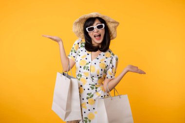 İlkbahar elbisesi, şapkası, güneş gözlüğü modası ve sarı arka planda izole edilmiş kese kağıdı olan çekici, mutlu Asyalı bir kadın. Yaz mevsiminde alışveriş konsepti.