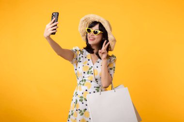Çekici, mutlu Asyalı bir kadın şık bir bahar elbisesi, şapkası, güneş gözlüğü modası ve sarı arka planda izole edilmiş kese kağıdıyla akıllı telefon kullanıyor. Yaz mevsiminde alışveriş konsepti.