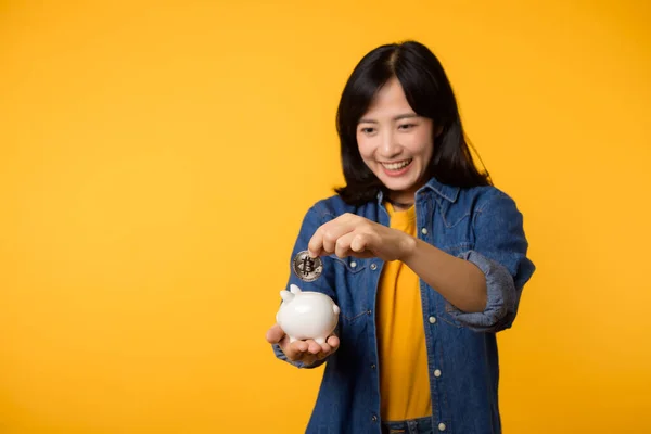 黄色のTシャツデニムシャツを着た幸せなアジアの若い女性は デジタル暗号通貨を黄色の背景に孤立した貯金箱に保存します デジタルマネー貯蓄投資の概念 — ストック写真
