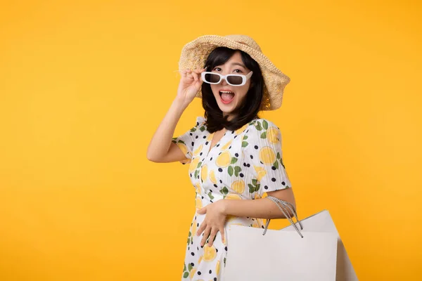 stock image Portrait attractive happy young asian woman with trendy springtime dress, hat, sunglasses fashion and paper bag isolated on yellow background. Summertime sale shopping concept.