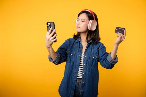 スマートフォンやクレジットカードを持ち デニムの服やヘッドフォンを身に着けている疑いの顔を持つ肖像アジアの若い女性 ショッピングオンライン携帯電話エンターテイメントライフスタイルコンセプト — ストック写真