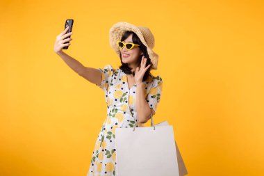 Çekici, mutlu Asyalı bir kadın şık bir bahar elbisesi, şapkası, güneş gözlüğü modası ve sarı arka planda izole edilmiş kese kağıdıyla akıllı telefon kullanıyor. Yaz mevsiminde alışveriş konsepti.