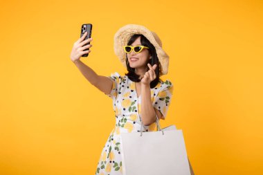 Çekici, mutlu Asyalı bir kadın şık bir bahar elbisesi, şapkası, güneş gözlüğü modası ve sarı arka planda izole edilmiş kese kağıdıyla akıllı telefon kullanıyor. Yaz mevsiminde alışveriş konsepti.