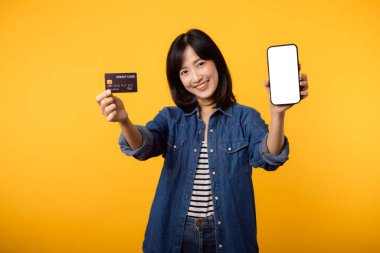 Güzel Asyalı genç bir kadının portresi. Kot ceket giymiş mutlu bir gülümseme. Stüdyonun sarı arka planında akıllı telefon ve kredi kartını gösteriyor. Çevrimiçi akıllı telefon uygulaması alışverişi