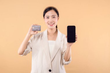 Beyaz ceketli mutlu Asyalı iş kadınının portresi. Elinde akıllı telefon ve kredi kartı bej arka planda duruyor. alışveriş kavramı.