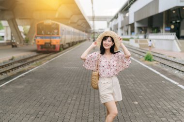 Sepet ören Asyalı genç kadın gezgin mutlu bir şekilde gülümsüyor ve arka planda trenin yanındaki kameraya bakıyor. Seyahat yaşam tarzı, dünya turisti ya da Asya yaz turizmi konsepti.