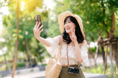 Yeşil park arka planında şapkası, sepeti, cep telefonu ve kamerası olan Asyalı genç bir kadın gezginin portresi. Seyahat yaşam tarzı, dünya turisti ya da Asya yaz turizmi konsepti.