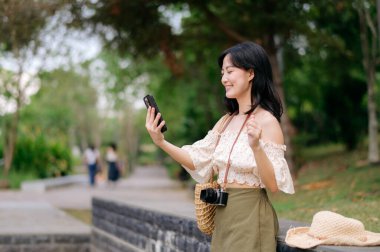 Yeşil park arka planında şapkası, sepeti, cep telefonu ve kamerası olan Asyalı genç bir kadın gezginin portresi. Seyahat yaşam tarzı, dünya turisti ya da Asya yaz turizmi konsepti.