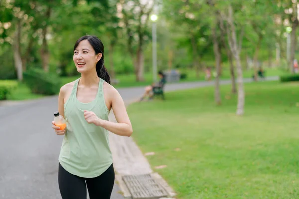 女慢跑者 让亚洲的年轻女子穿着绿色运动服 跑步后喝着有机橙汁 享受着健康的户外生活 健身跑步的女孩在公共公园 健康是概念 — 图库照片