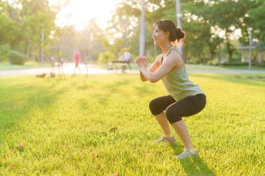 Kadın koşucu. Formda, yeşil spor kıyafetli genç bir Asyalı kadın koşmadan ve dışarıda sağlıklı bir şekilde eğlenmeden önce parkta çömeliyor. Kamu parkındaki spor koşucusu kız. Refah konsept demek.