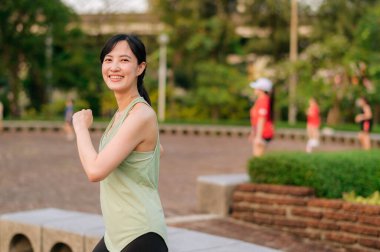 Kadın koşucu. Yeşil spor giyim aerobiği olan formda bir Asyalı genç kadın parkta dans ediyor ve açık havada eğleniyor. Kamu parkındaki spor koşucusu kız. Refah konsept demek.