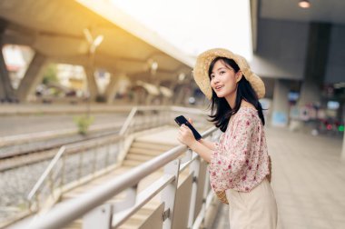 Bangkok 'taki tren istasyonunun yanında cep telefonu kullanarak dokuma sepetli genç Asyalı kadın gezgin. Seyahat yaşam tarzı, dünya turisti ya da Asya yaz turizmi konsepti.
