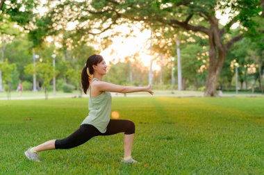 Kadın koşucu. Açık havada koşup eğlenmeden önce yeşil spor kıyafetli sağlıklı bir Asyalı genç kadın parkta kaslarını esnetiyor. Kamu parkındaki spor koşucusu kız. Refah konsept demek.