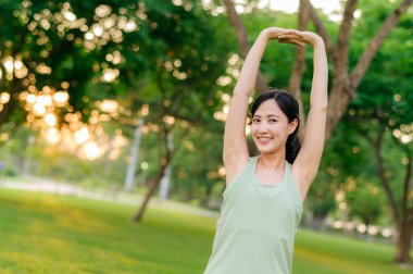 Kadın koşucu. Açık havada koşup eğlenmeden önce yeşil spor kıyafetli sağlıklı bir Asyalı genç kadın parkta kaslarını esnetiyor. Kamu parkındaki spor koşucusu kız. Refah konsept demek.