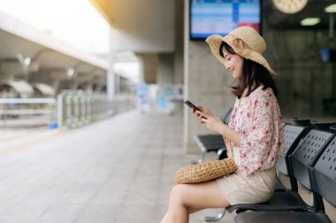 Cep telefonu kullanıp tren istasyonunda bekleyen sepetli Asyalı genç bir kadın gezgin. Seyahat yaşam tarzı, dünya turisti ya da Asya yaz turizmi konsepti.