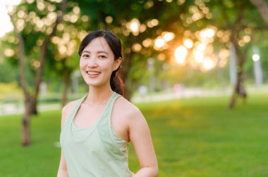 Kadın koşucu. Açık havada koşup eğlenmeden önce yeşil spor kıyafetli sağlıklı bir Asyalı genç kadın parkta kaslarını esnetiyor. Kamu parkındaki spor koşucusu kız. Refah konsept demek.