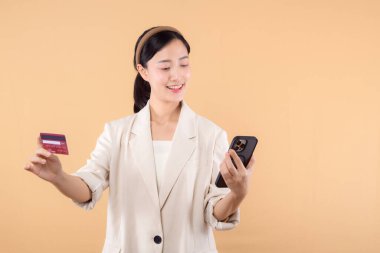 Beyaz ceketli mutlu Asyalı iş kadınının portresi. Elinde akıllı telefon ve kredi kartı bej arka planda duruyor. alışveriş kavramı.