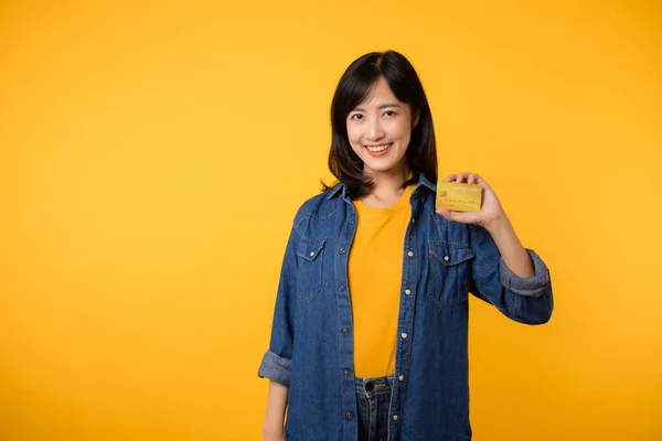 黄色のTシャツとデニムのジャケットを着たアジアの若い女性の肖像画で 幸せな笑顔が黄色の背景に孤立したクレジットカードを保持しています 支払いショッピングオンラインコンセプト — ストック写真